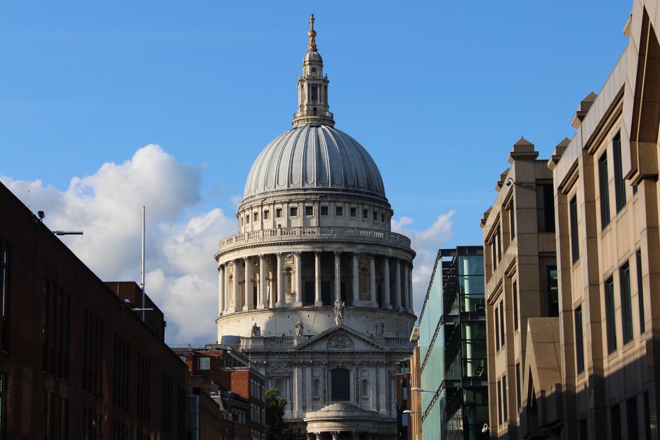 A city street scene featuring a large historic dome-shaped building with classical architectural elements such as columns and decorative sculptures, situated behind modern office buildings with rectan